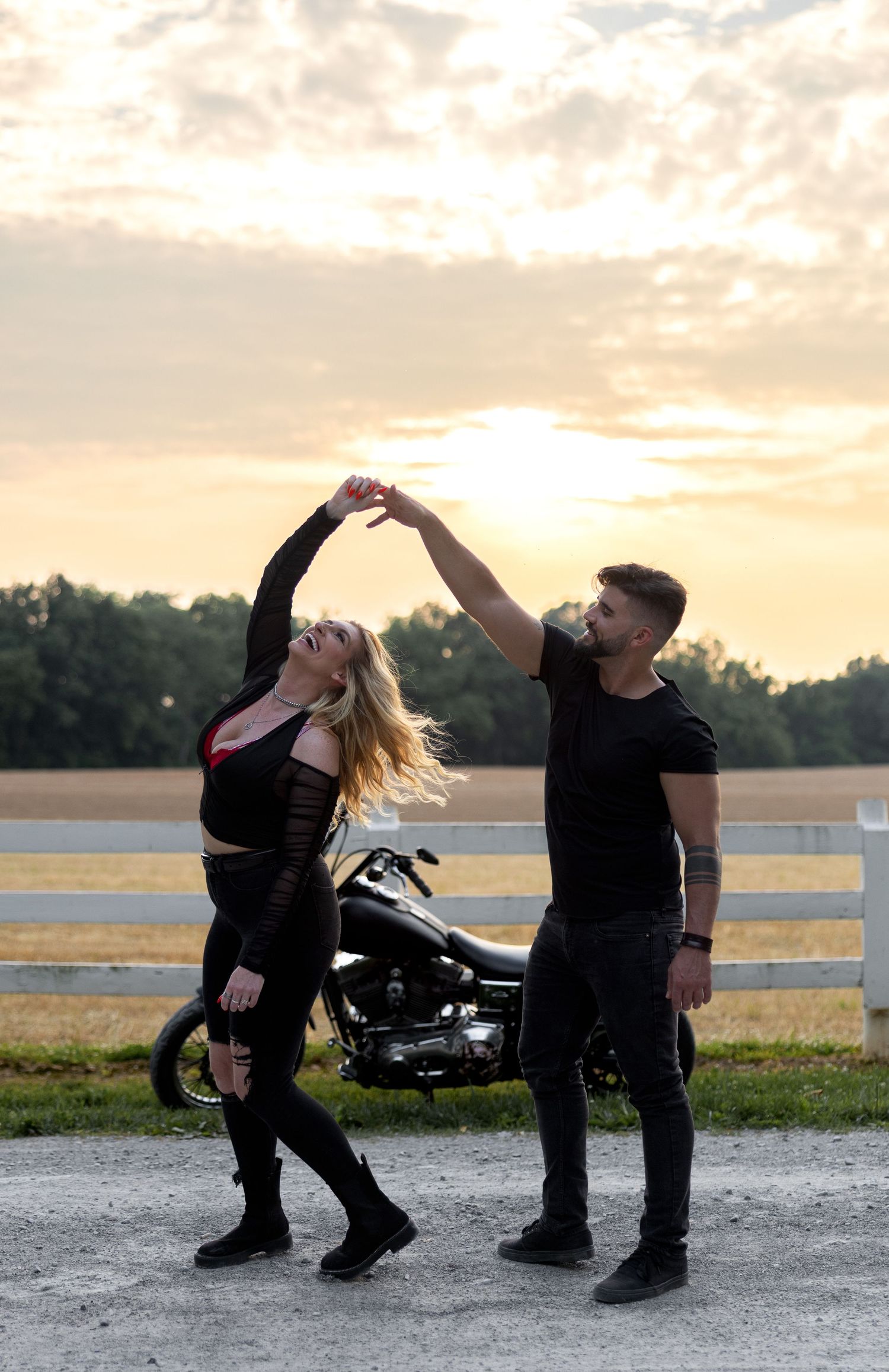 A romantic couple dancing together at sunset near a motorcycle and white fence.