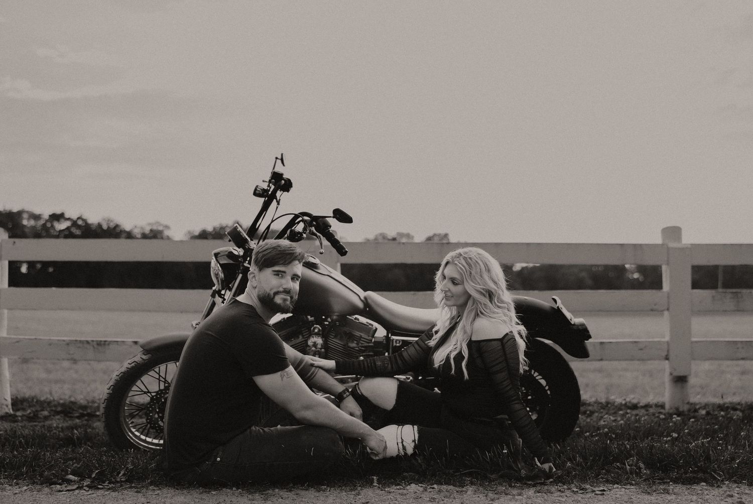 A black and white photo of a couple relaxing on grass beside a motorcycle and fence.