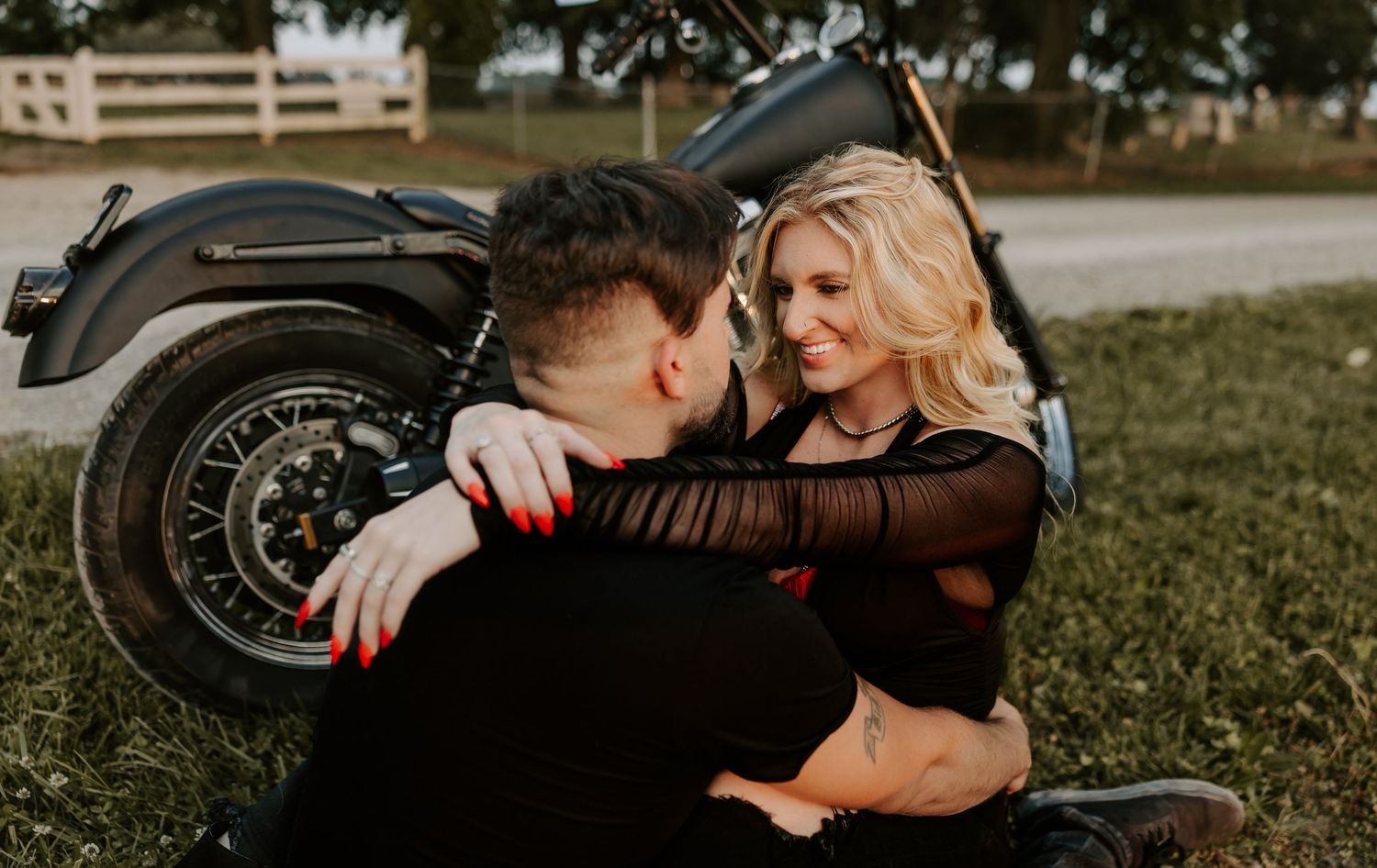 A couple sharing an intimate moment while sitting next to a motorcycle on grass.