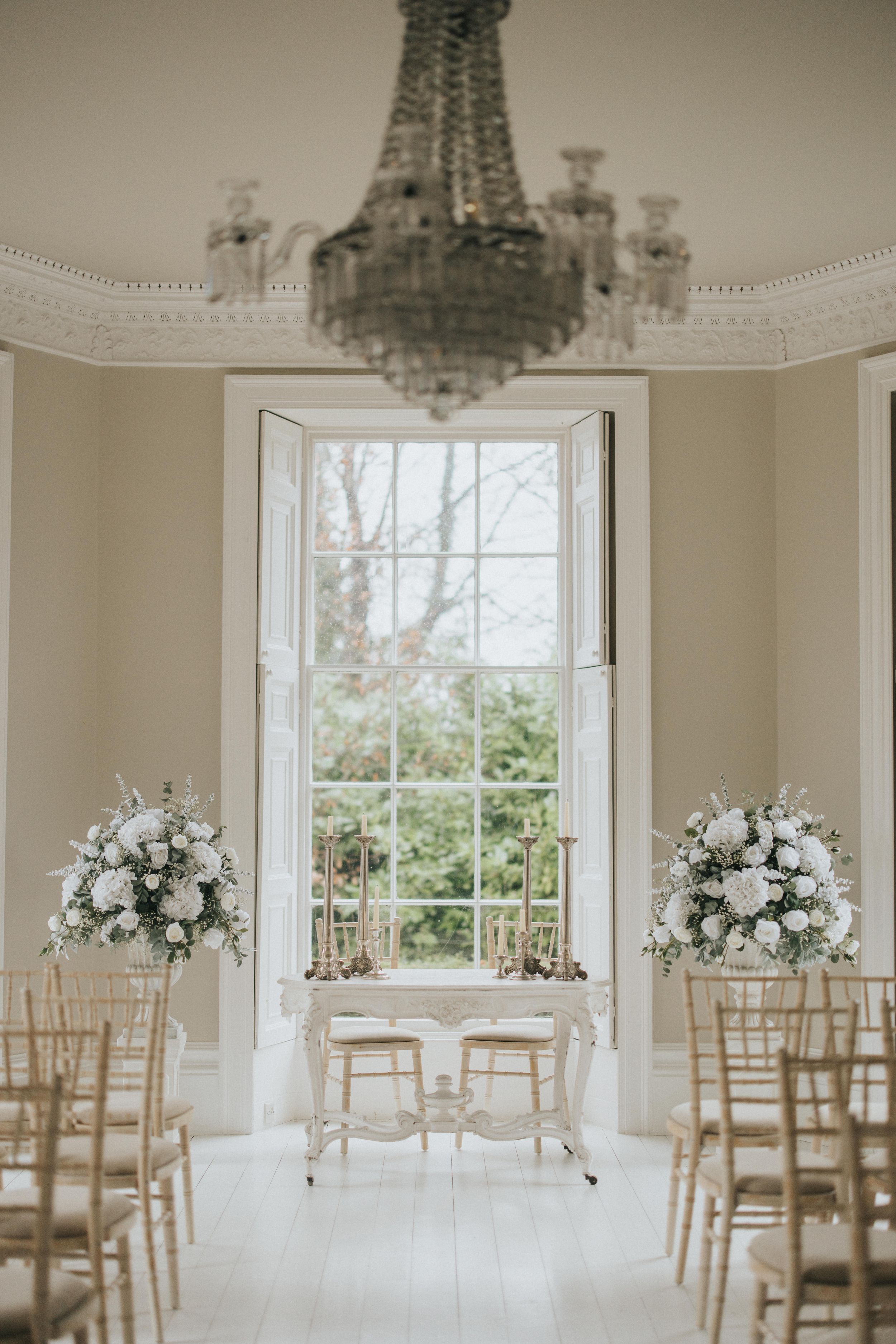 Elegant white wedding reception room with crystal chandelier, flower arrangements and wooden chiavari chairs in natural light.