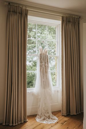 A delicate lace wedding dress hangs in front of a sunlit window with floor-length beige curtains in an elegant room.