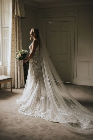 Moody bridal portrait series showing dramatic cathedral veil trailing across beige carpet in window light.