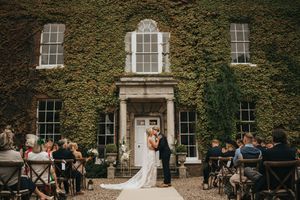 A wedding ceremony takes place at the entrance of an ivy-covered historic mansion with guests seated on both sides.