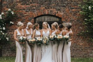 Bridesmaids wearing neutral champagne and beige dresses gather in front of rustic brick wall holding floral bouquets.