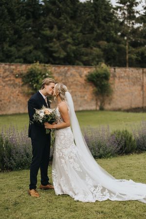 Happy couple shares intimate moment on their wedding day in a garden with lavender flowers and brick wall backdrop.