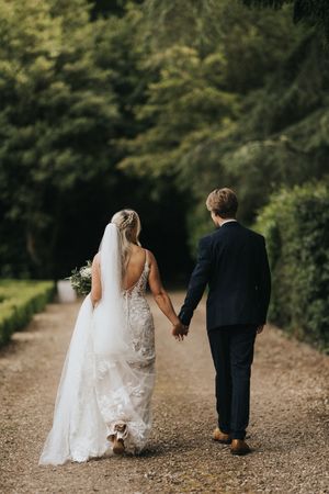 A wedding couple walks hand in hand down a gravel path bordered by lush green trees on a sunny day.