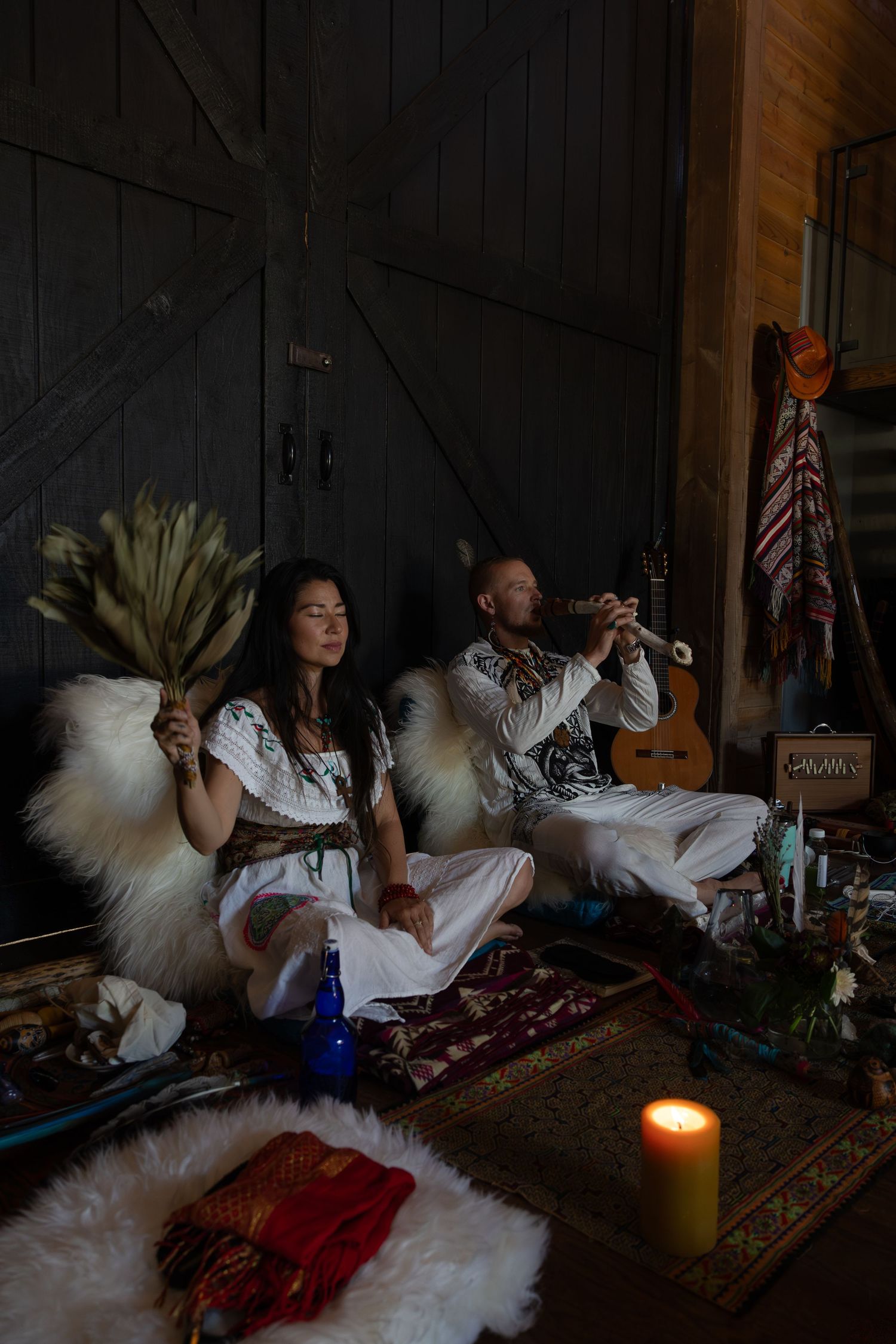 Two people in white outfits sit on furs with instruments and candles in a moody, bohemian indoor setting. A sacred ceremonial space.