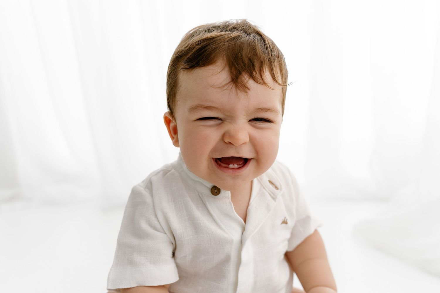 A joyful child wearing a white button-down shirt laughs expressively against a bright white background.