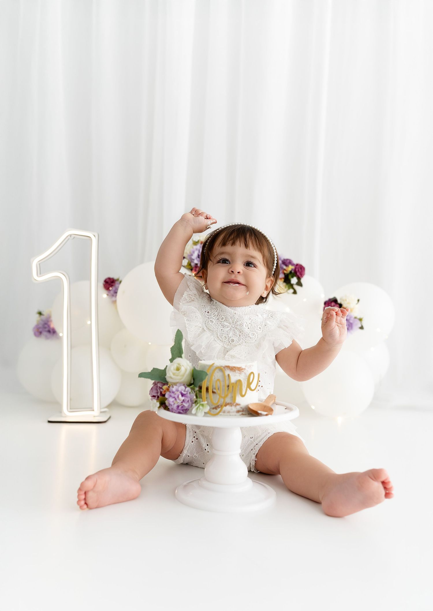 A baby sits next to a glowing number one and cake stand with flowers during a first birthday photoshoot.