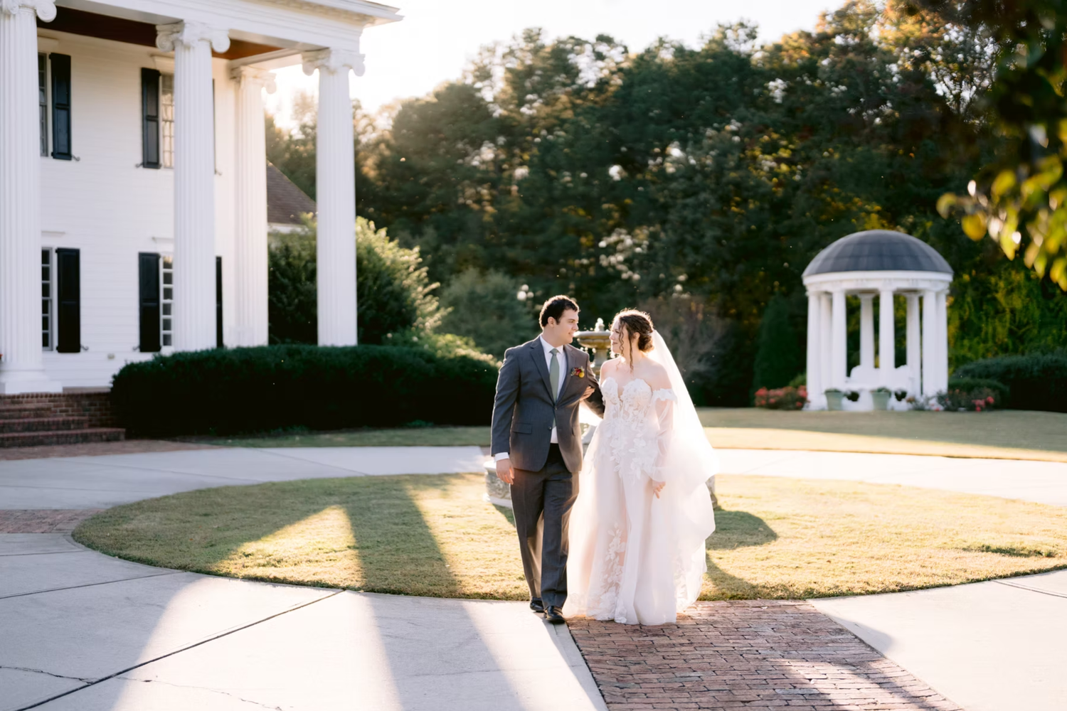 Couple in wedding attire walk along pathway at elegant white mansion with gazebo in late afternoon sunlight.