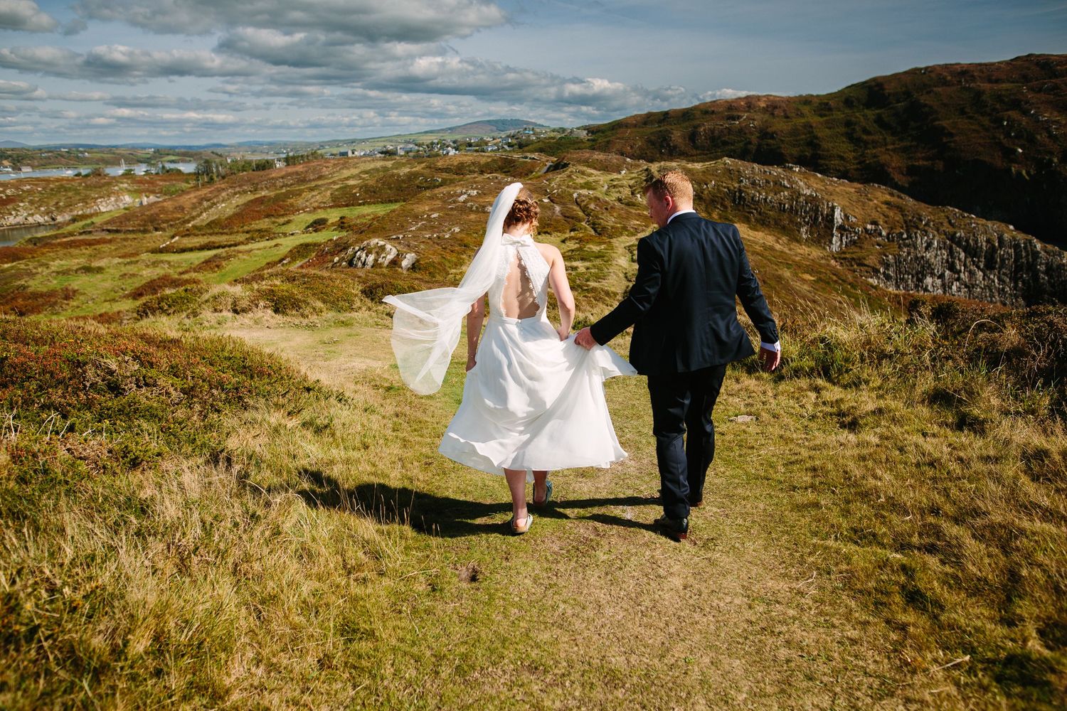 A couple in wedding attire walks hand in hand along a scenic hilltop path with rolling green landscapes and cloudy skies.