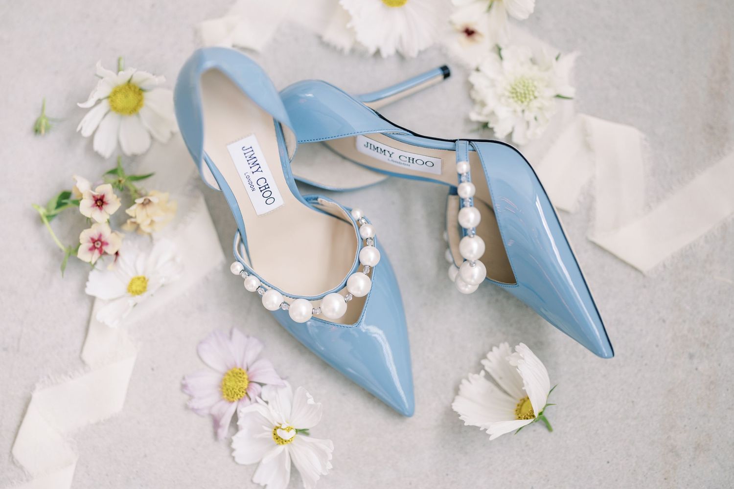 Light blue pointy toe heels surrounded by delicate white daisies and pearl accessories on a soft gray background.