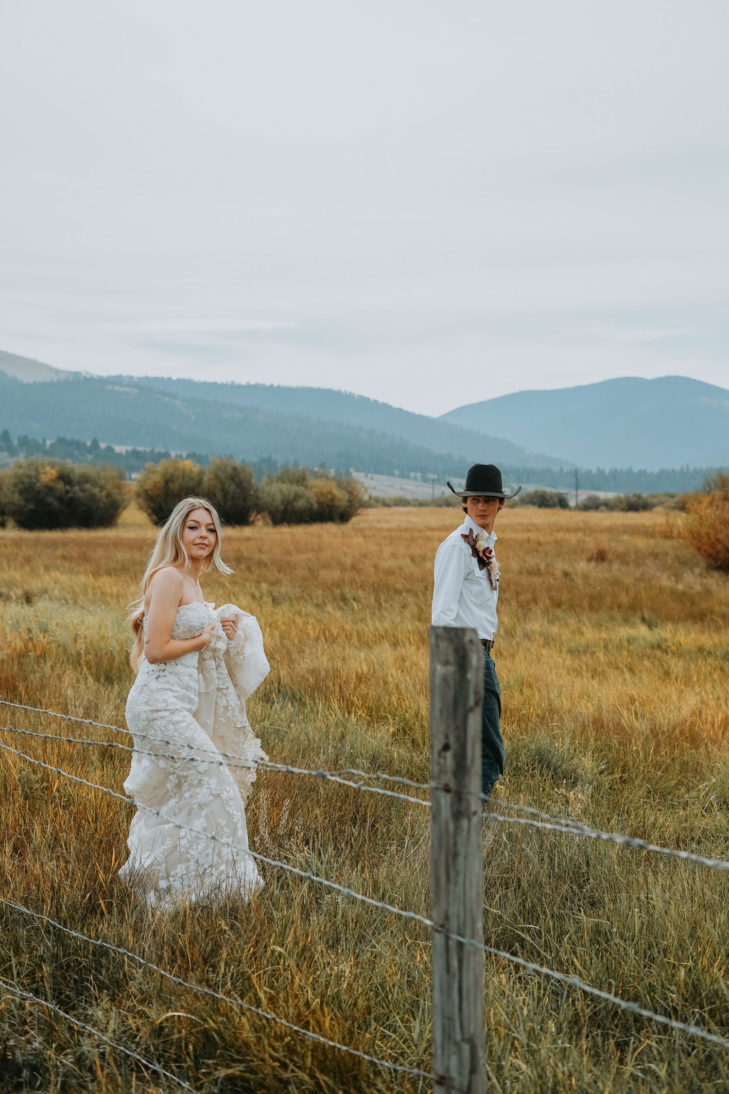 A couple stands in a meadow overlooking rolling hills and mountains in a moody black and white landscape photograph.