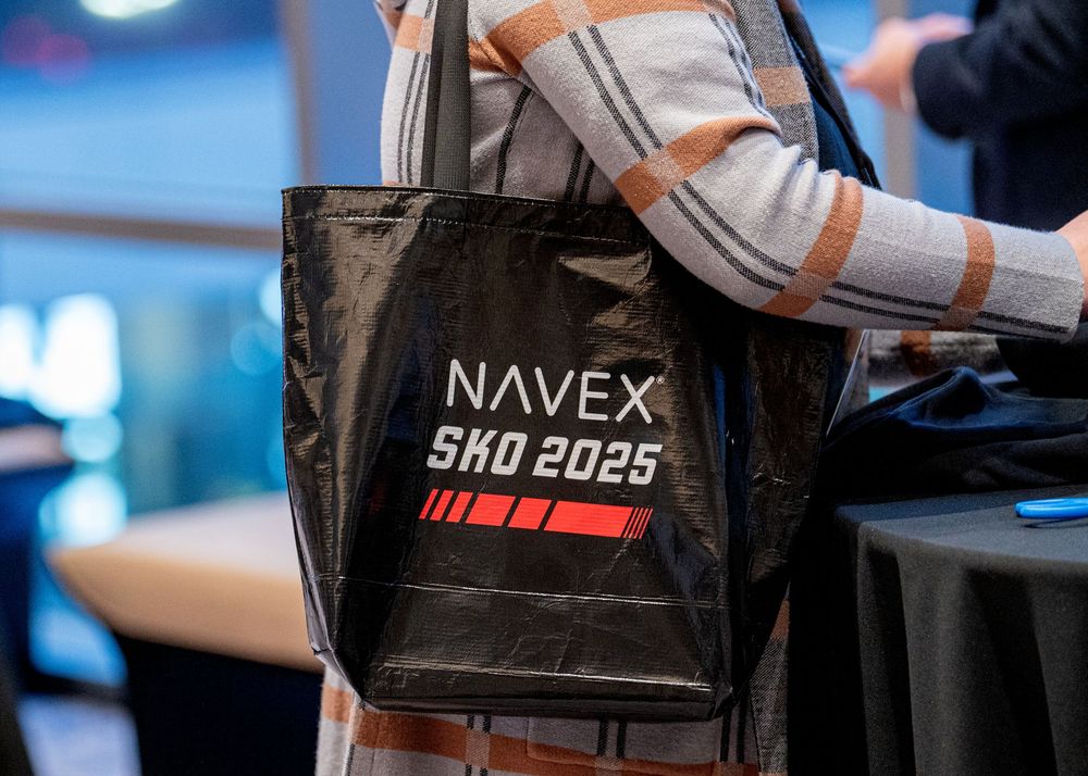 Black promotional tote bag featuring NAVEX SHO 2025 branding being carried at an event.