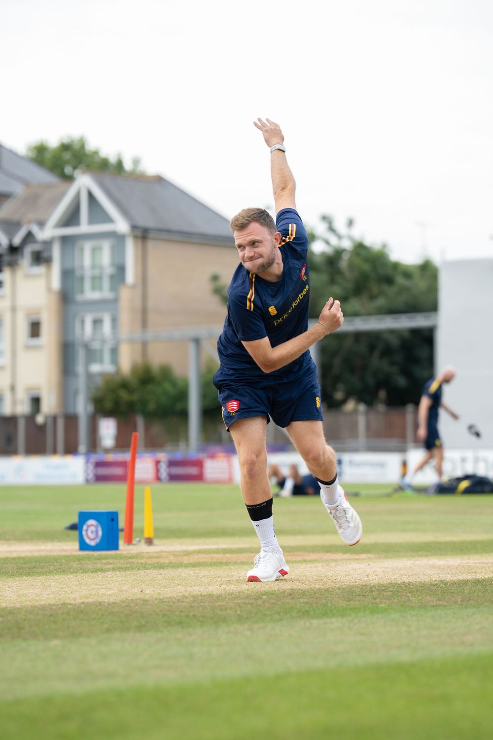 Cricket bowler in navy blue uniform mid-delivery stride on cricket pitch during daytime match.