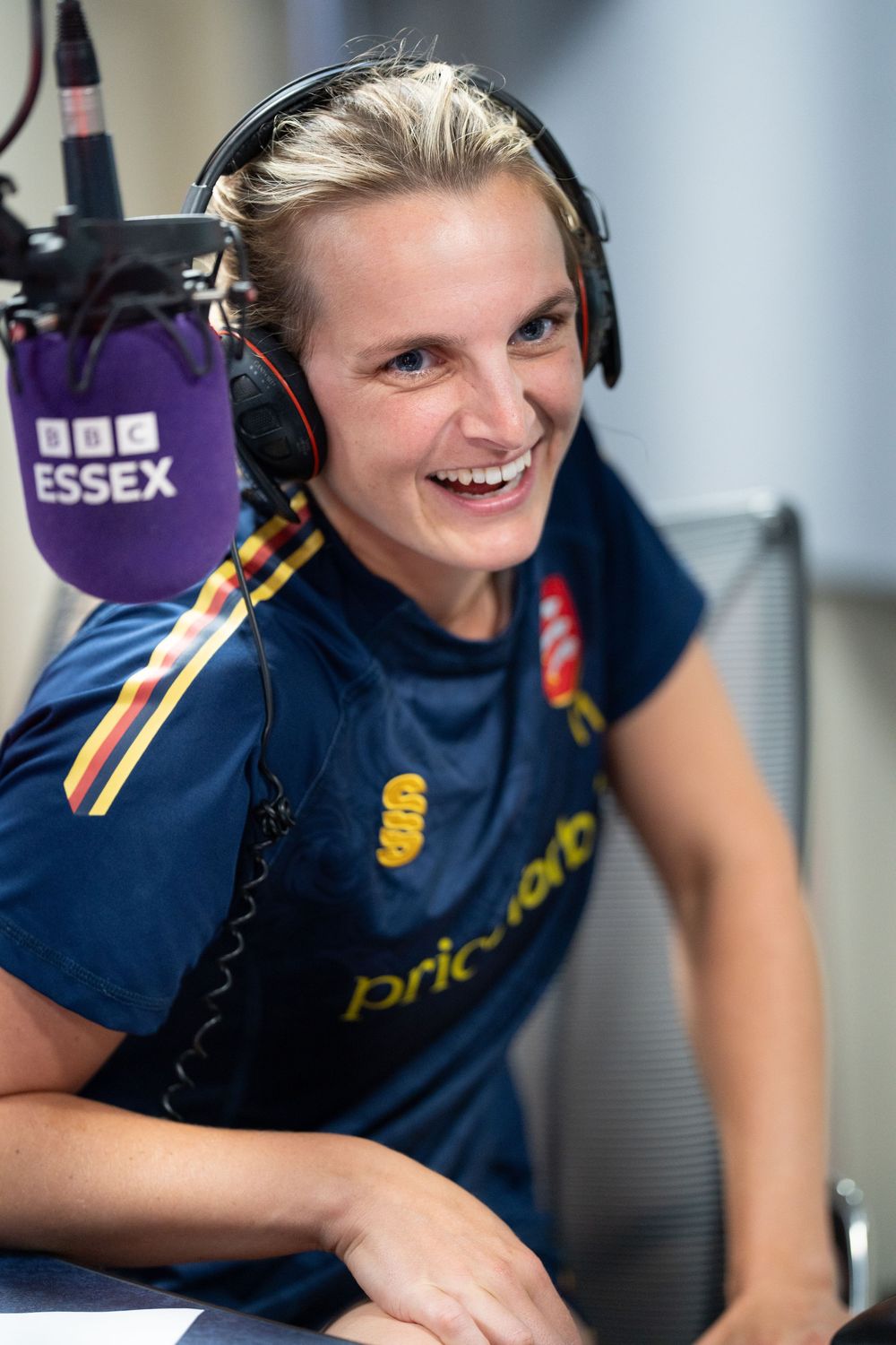 A radio host wearing headphones smiles while broadcasting at BBC Essex wearing a navy colored sports uniform.