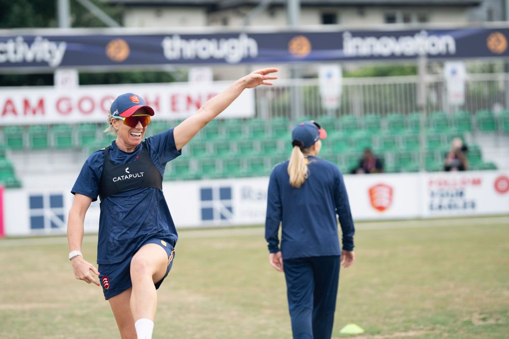 Cricket players in navy blue uniforms practicing on a sports field with stadium seating visible in the background.