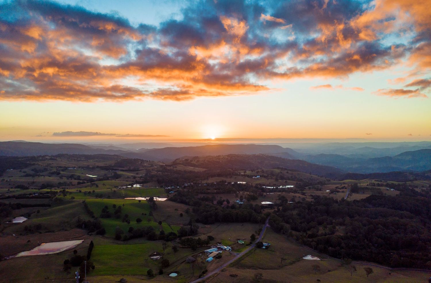 Rural sunset landscape with rolling hills and scattered buildings beneath dramatic orange clouds.