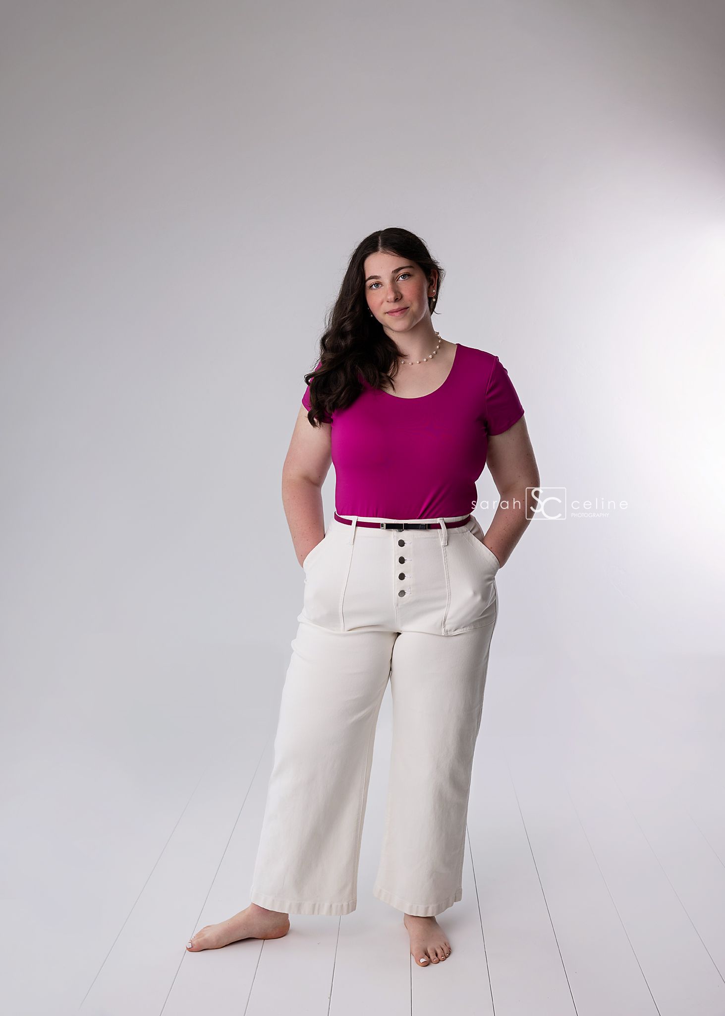 Woman in magenta t-shirt and high-waisted cream wide-leg pants stands barefoot against white background.