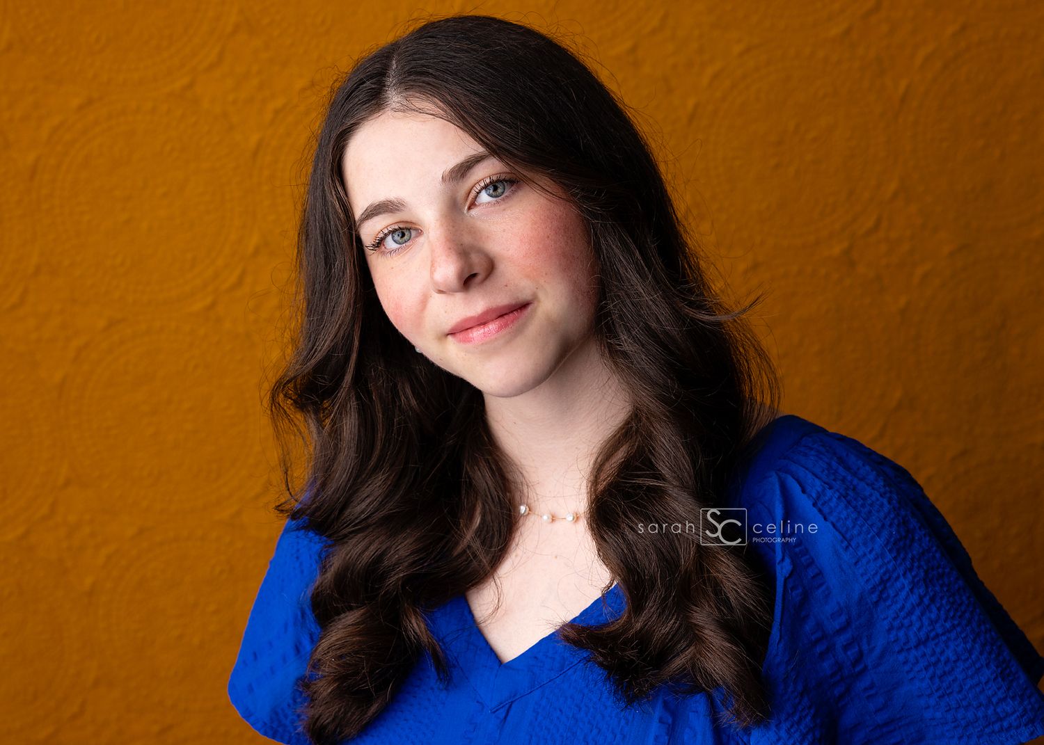 Portrait of someone in a blue v-neck top against an orange background with long dark wavy hair.
