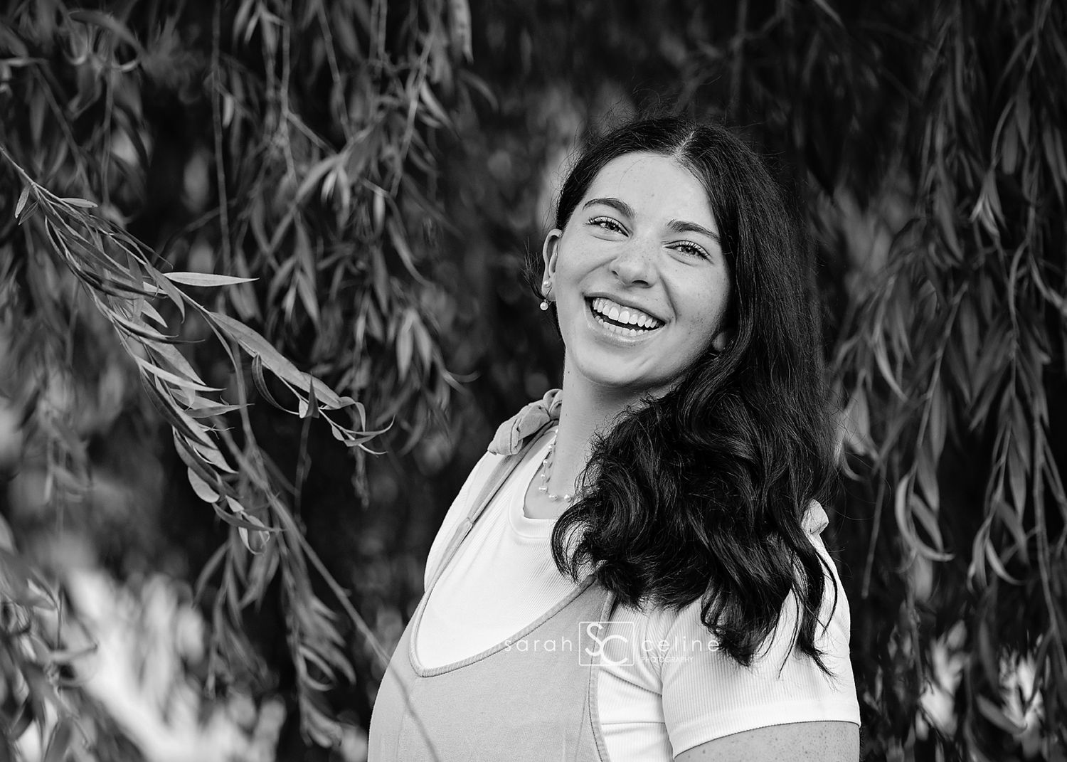 A woman with dark wavy hair sits among willow branches in a black and white outdoor portrait.