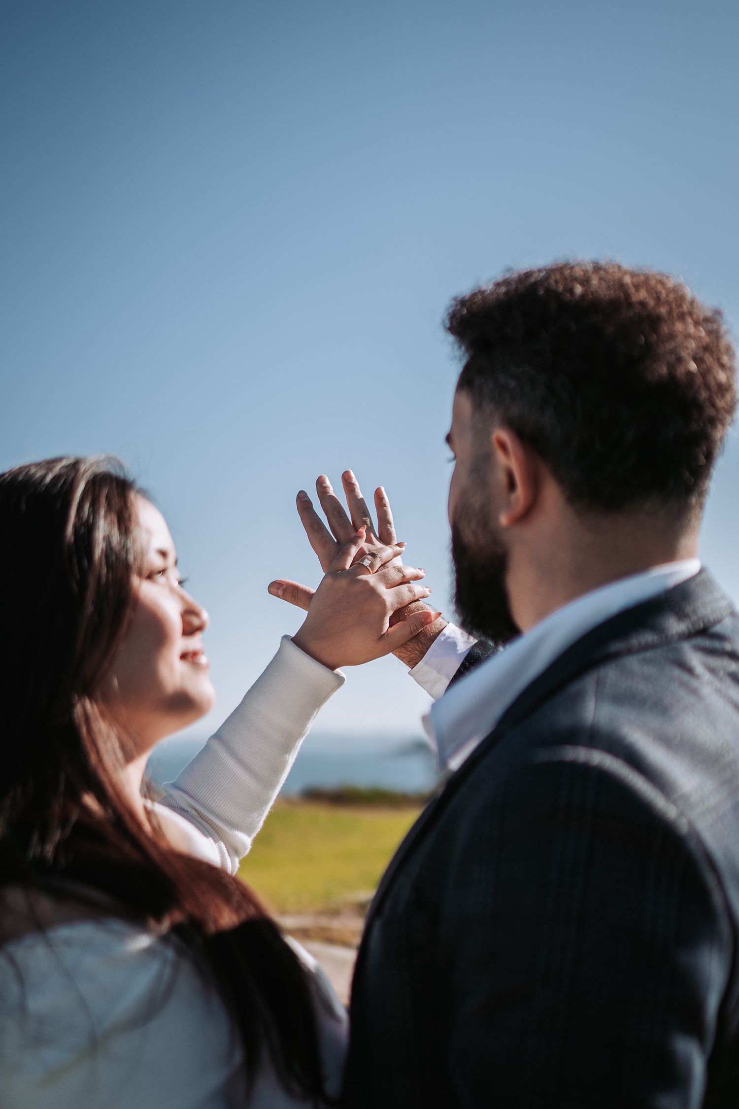 Couple showing off engagement ring outdoors against blue sky and sunny field backdrop.