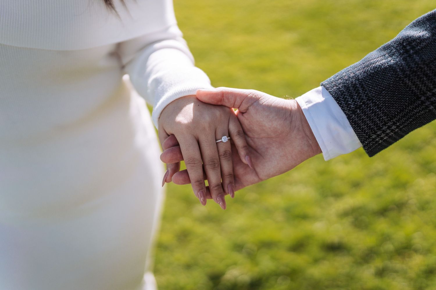 A close-up view of a couple holding hands lovingly while wearing bridal attire in front of a green lawn background.