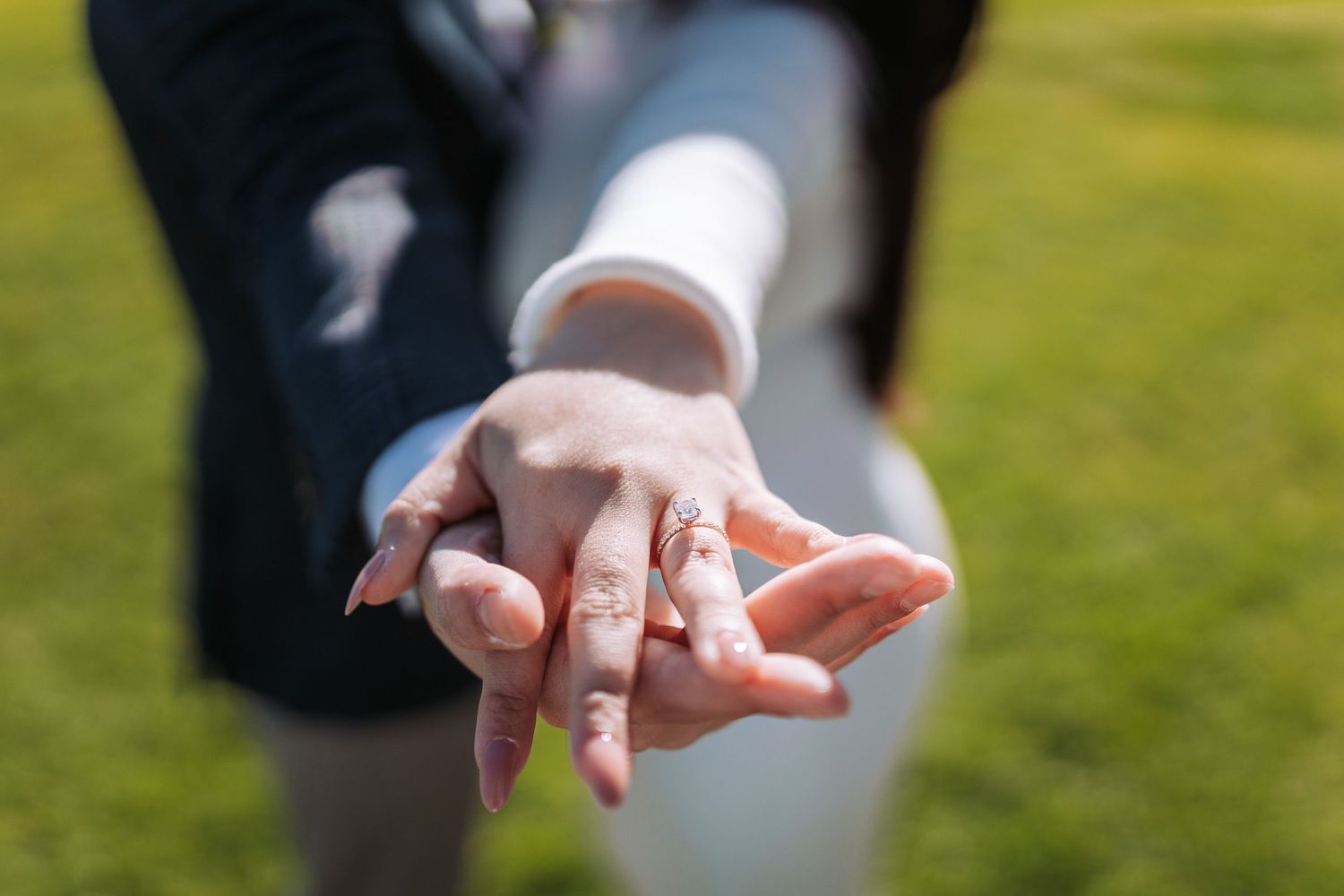 A hand holds out a small silver ring with a diamond solitaire against a blurred green outdoor background.