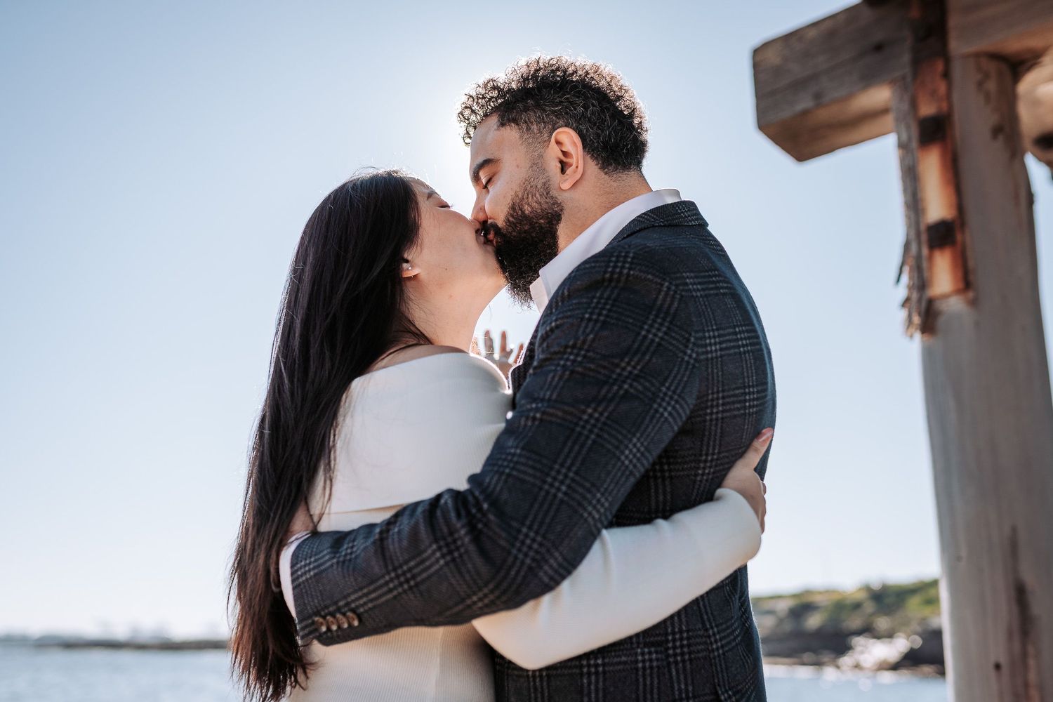 A couple sharing a romantic kiss under wooden beams with scenic water and coastline views in the background.