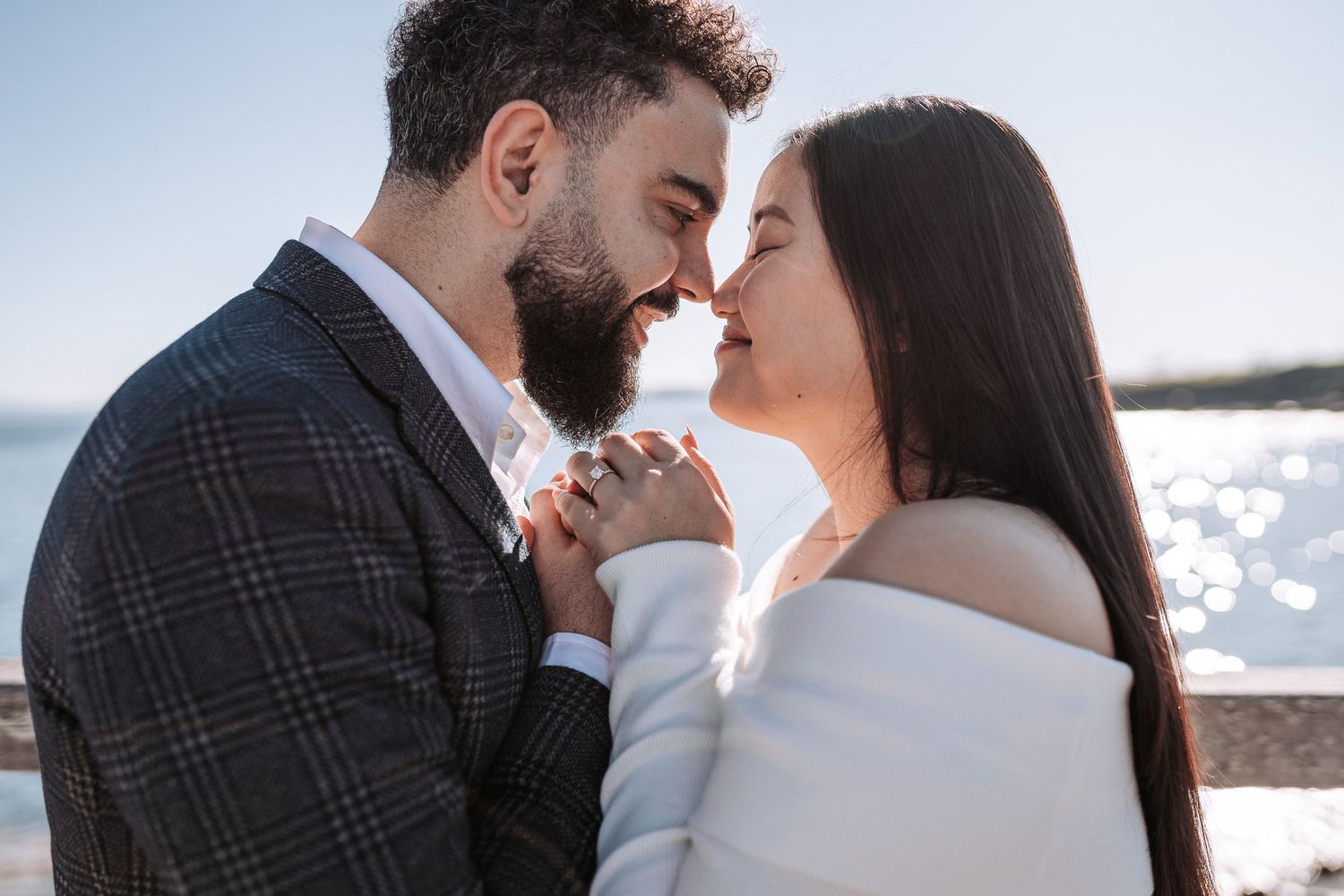Romantic couple in intimate pose share tender moment by sparkling waterfront on sunny day.