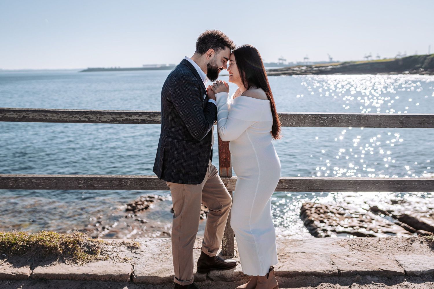 Couple embracing on rocky coastal shoreline with gentle waves and blue water in background.