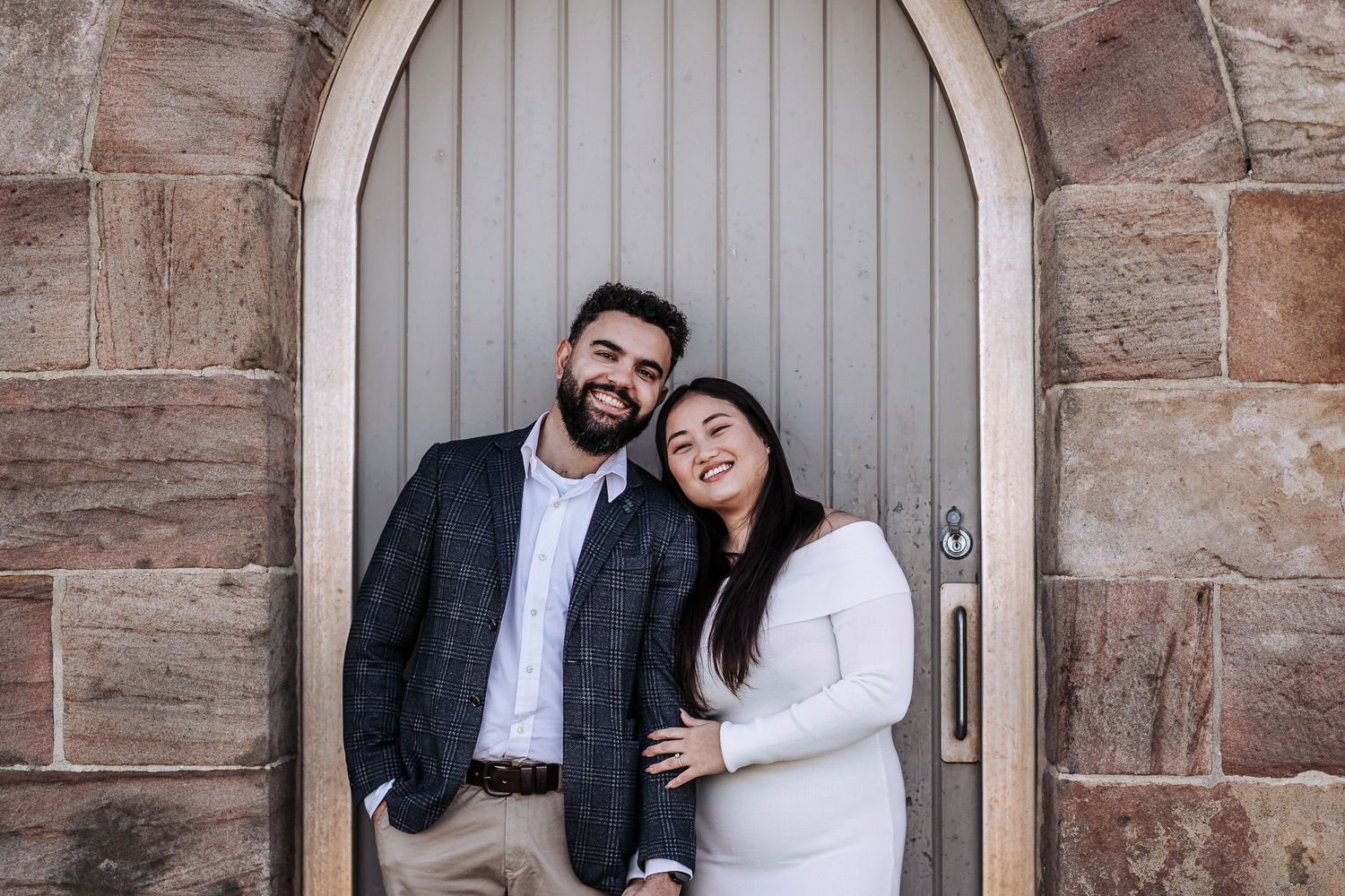 A happy couple stands together in front of an arched wooden door with stone walls surrounding it.