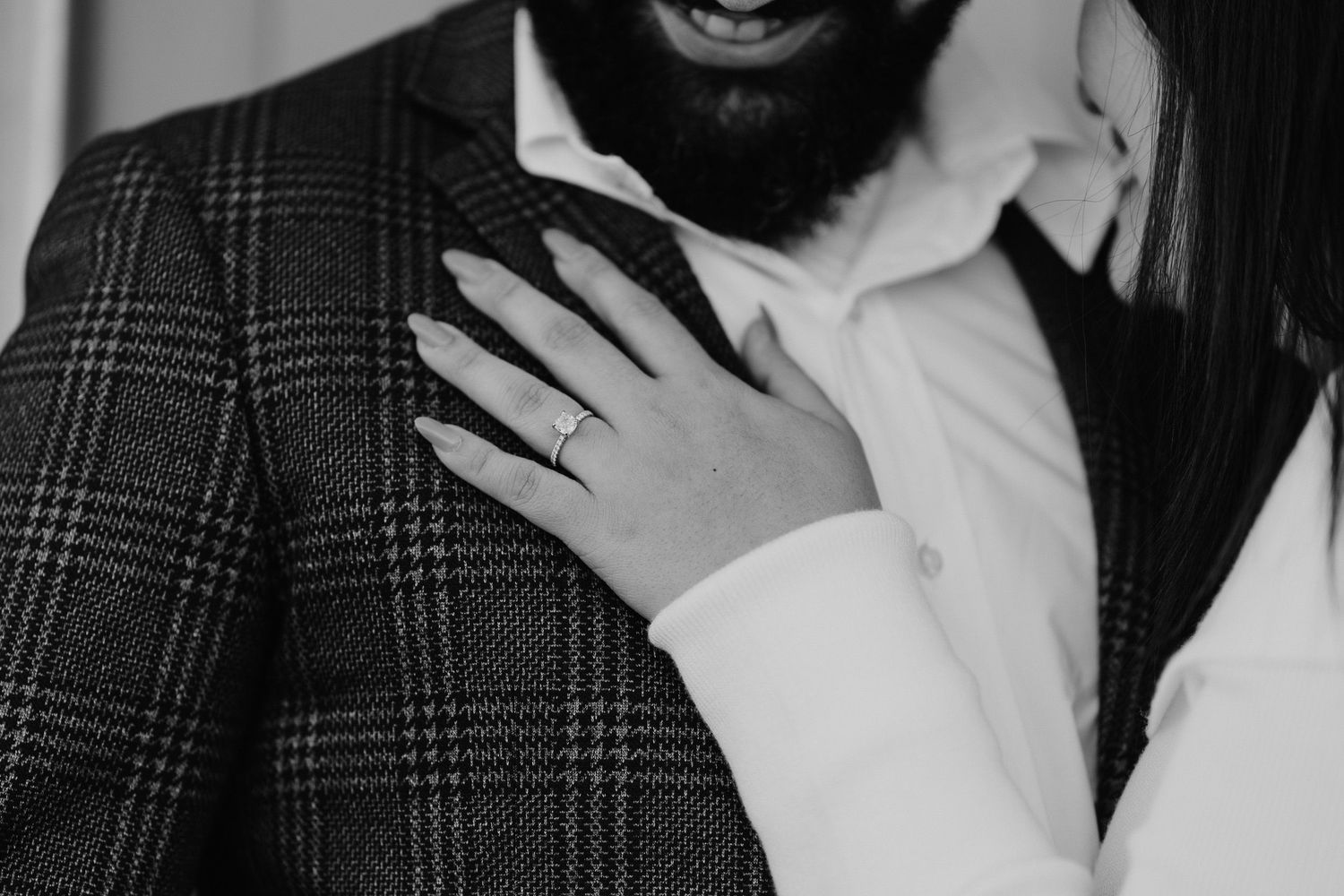 Black and white photo showing a hand with an engagement ring resting on a plaid suit jacket lapel.
