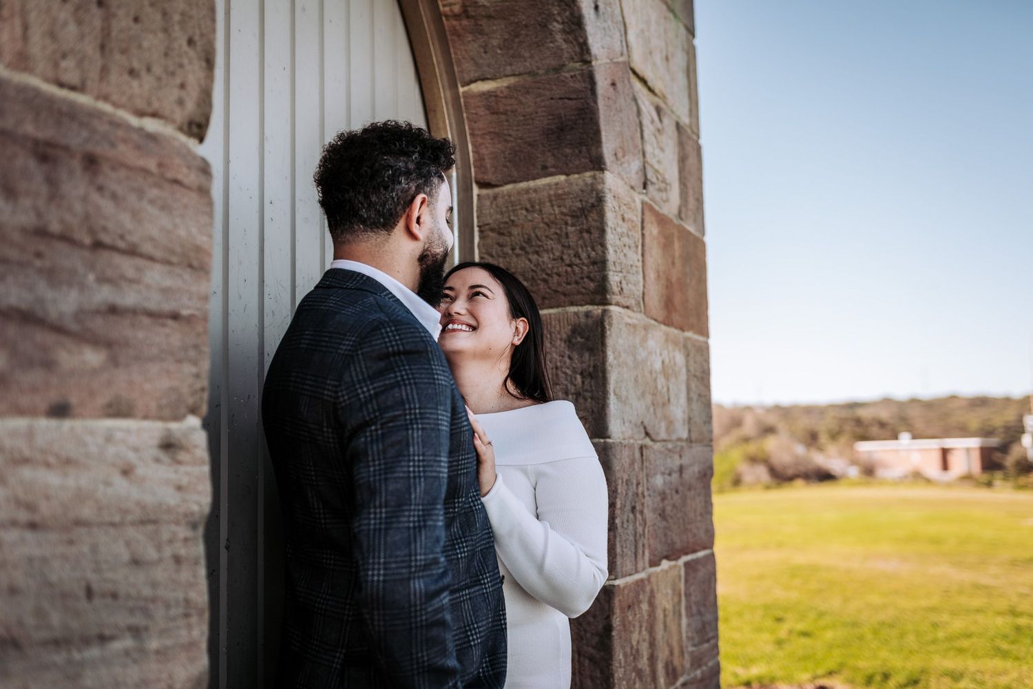 A tender embrace between a couple against a rustic stone wall with a green countryside in the background.
