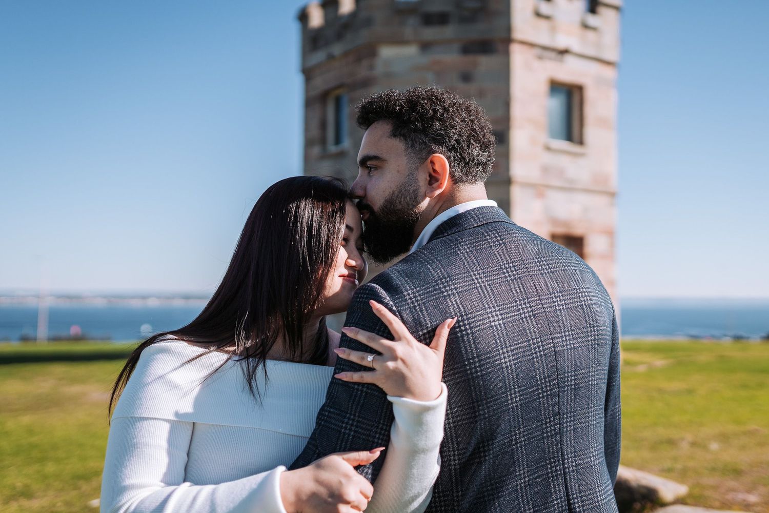 A loving couple sharing an intimate moment near a lighthouse overlooking the ocean on a sunny day.