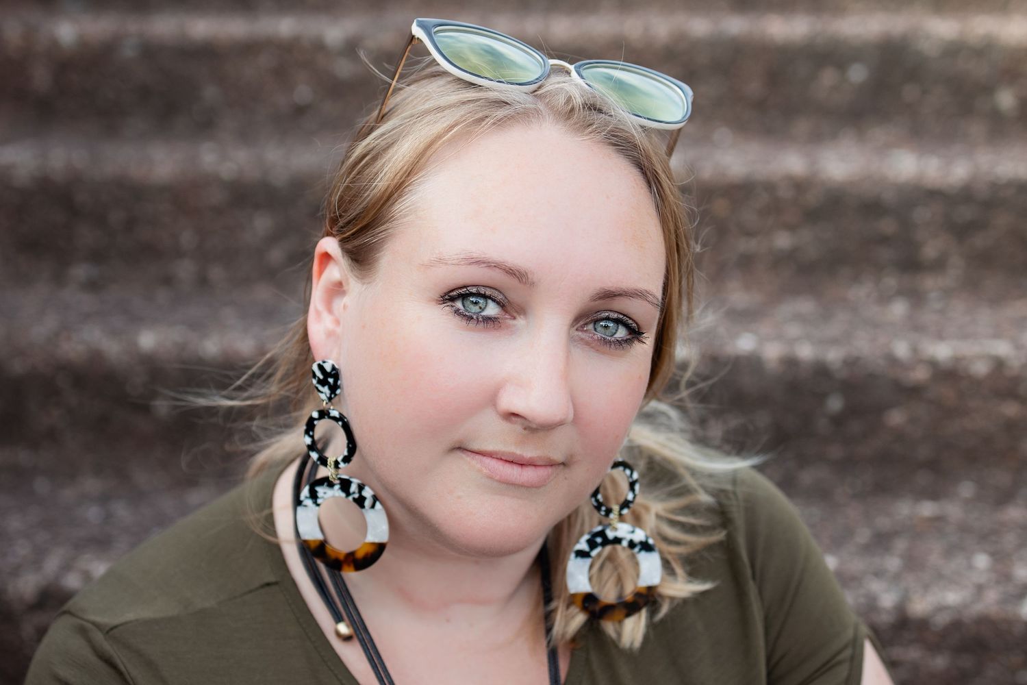 Outdoor close up portrait of woman sitting on concrete steps. She wears large earrings and sunglasses on her head.