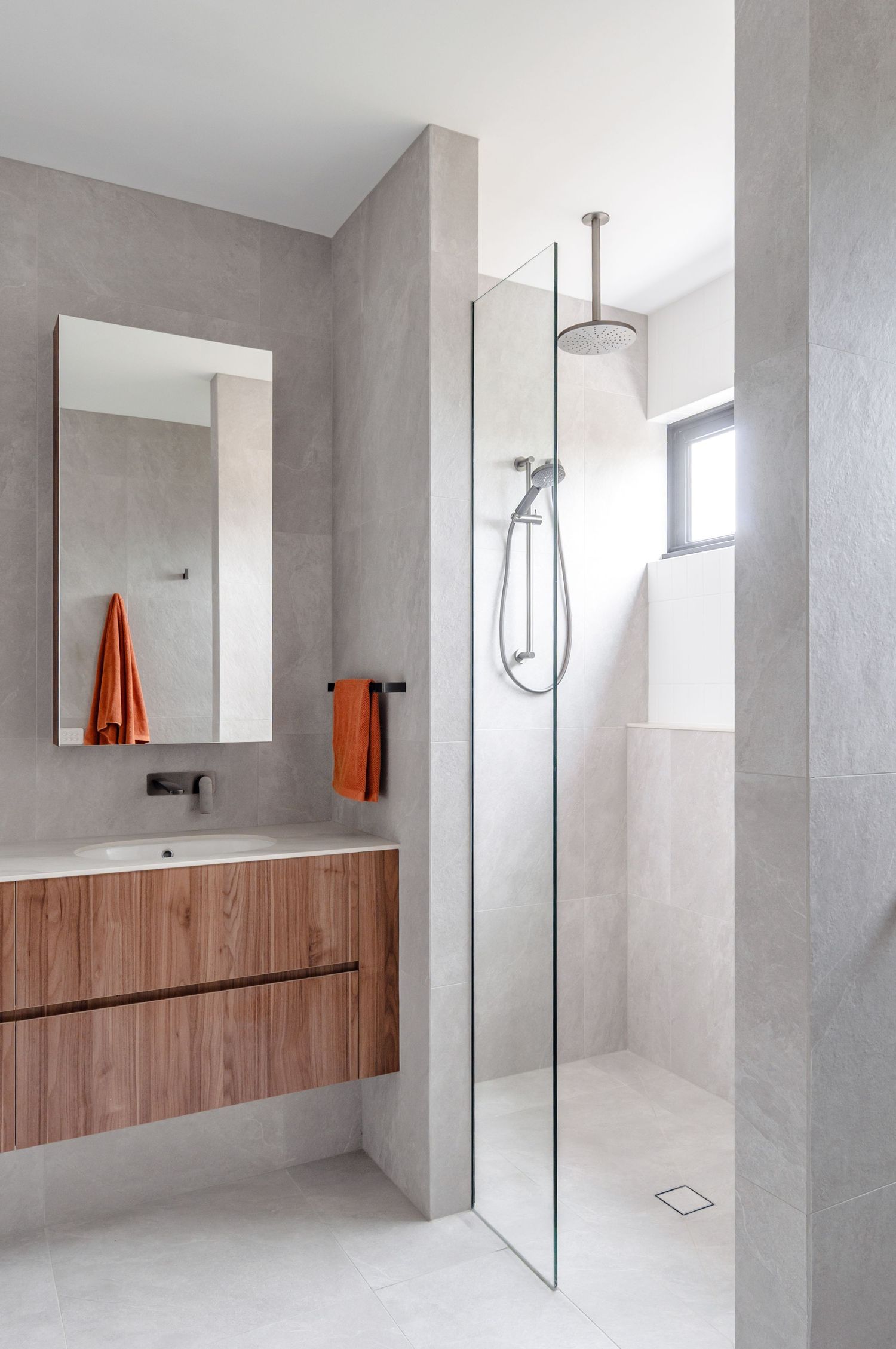 Contemporary bathroom with floating wood vanity, grey walls, glass shower partition and orange towel accent.