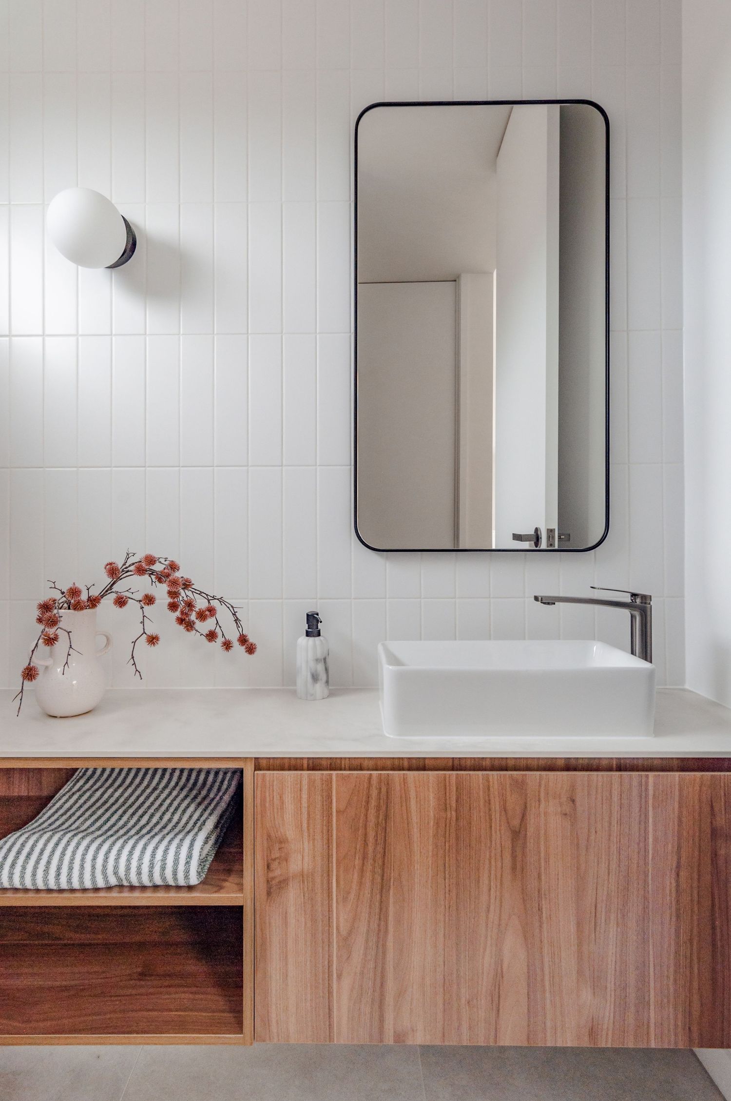 Modern bathroom vanity with wooden cabinets, white vessel sink, black framed mirror and decorative red branches.