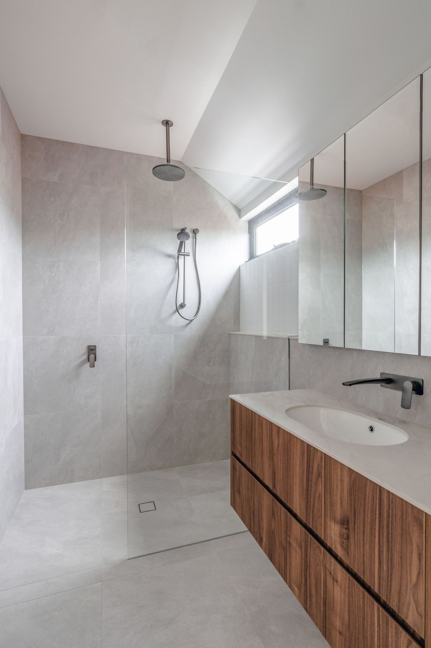 Contemporary bathroom featuring walnut vanity, walk-in shower with rainfall showerhead and light gray tile walls.