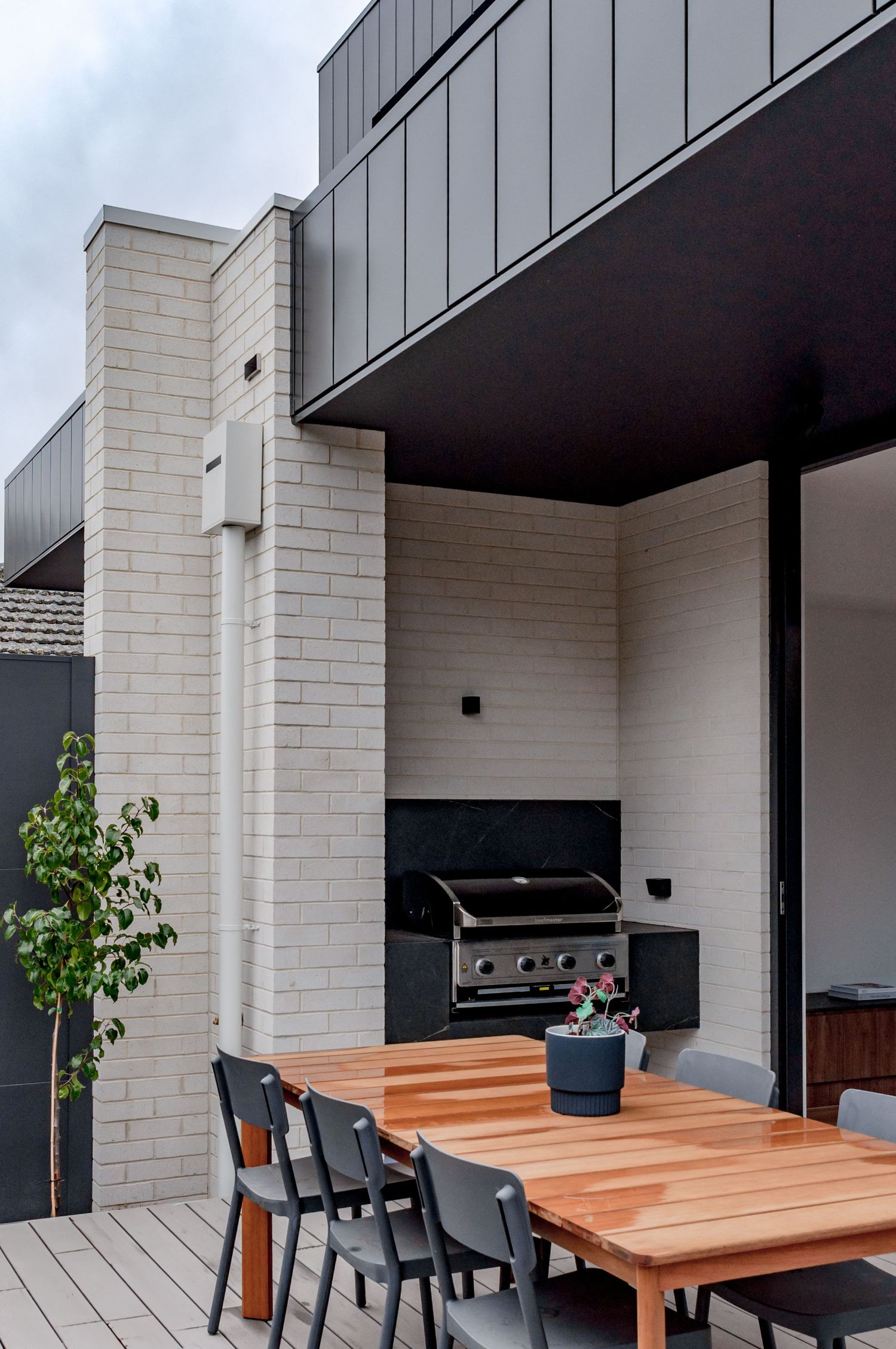 Modern outdoor patio space with wooden dining table and built-in barbecue against white brick walls.