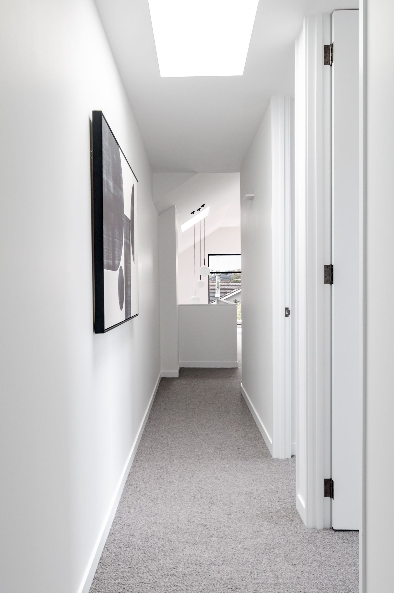 Modern minimalist hallway with white walls, grey carpet, black framed artwork and natural light from skylight.