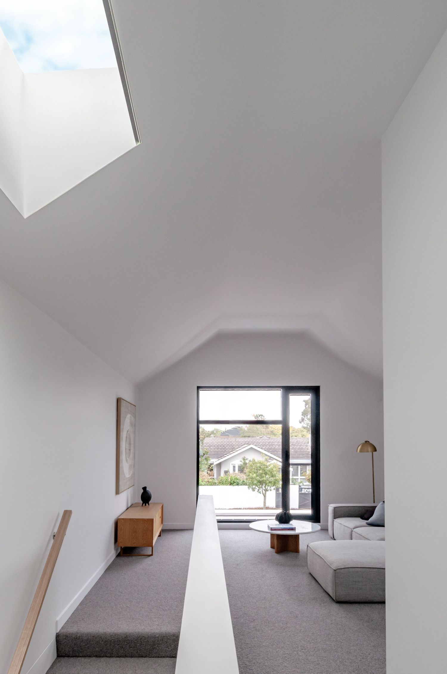 Modern attic bedroom with sloped white ceiling, grey carpet, and black-framed window creating a minimalist space.