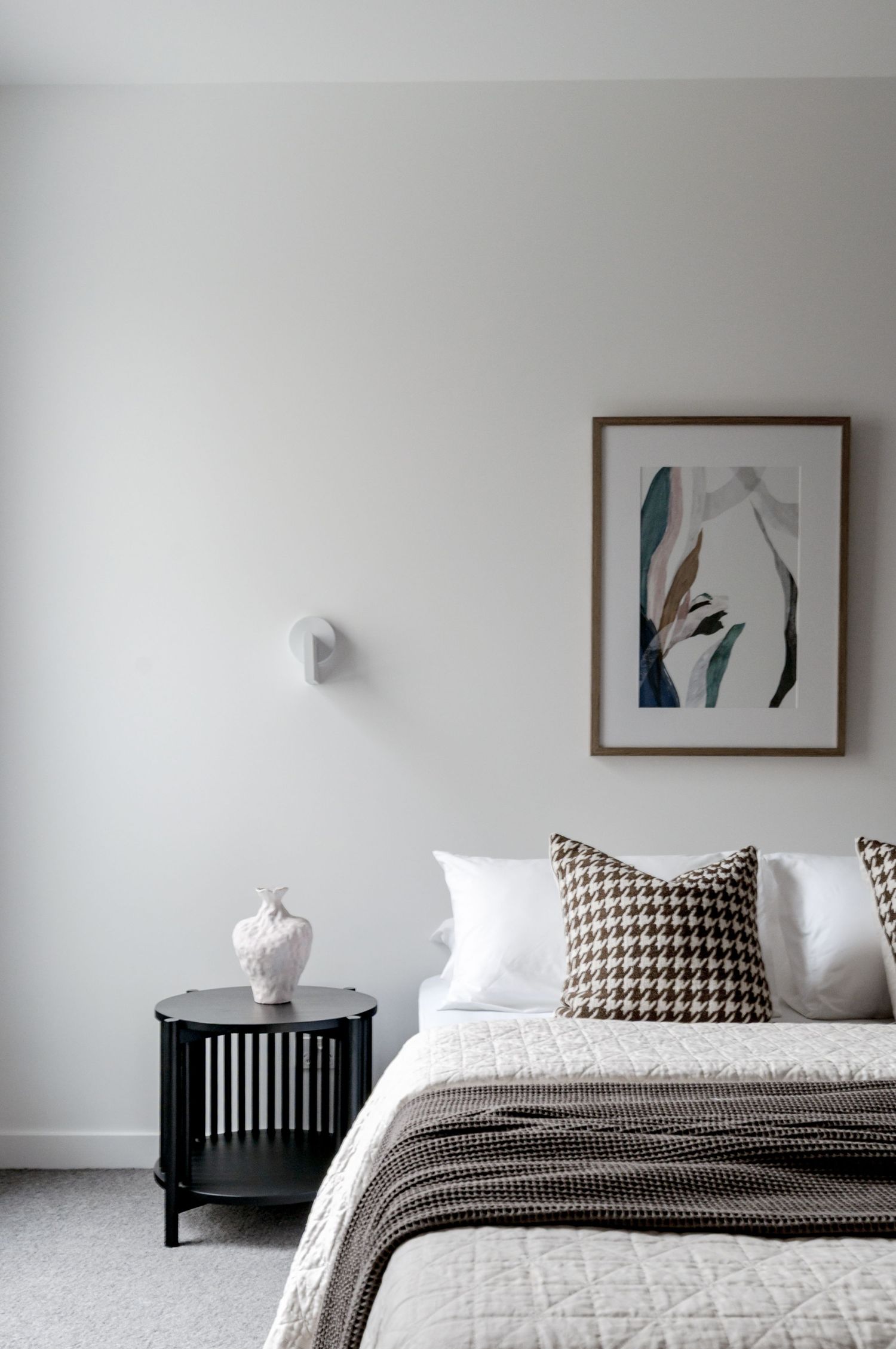 Minimalist bedroom with black nightstand, houndstooth pillow, and abstract wall art in neutral tones.