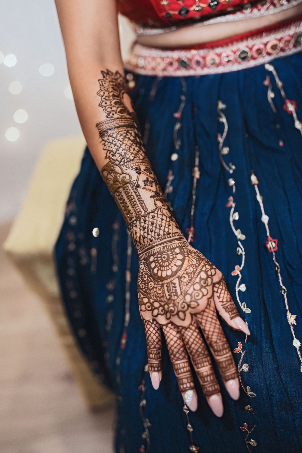 Intricate henna mehndi design adorns hand and arm against dark blue traditional dress with embellishments.