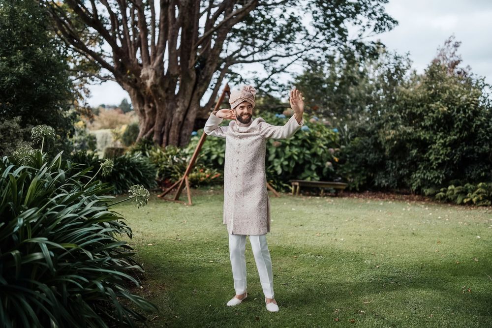 Person in light colored clothing stands with raised arms in a lush garden setting with large trees and plants in background.