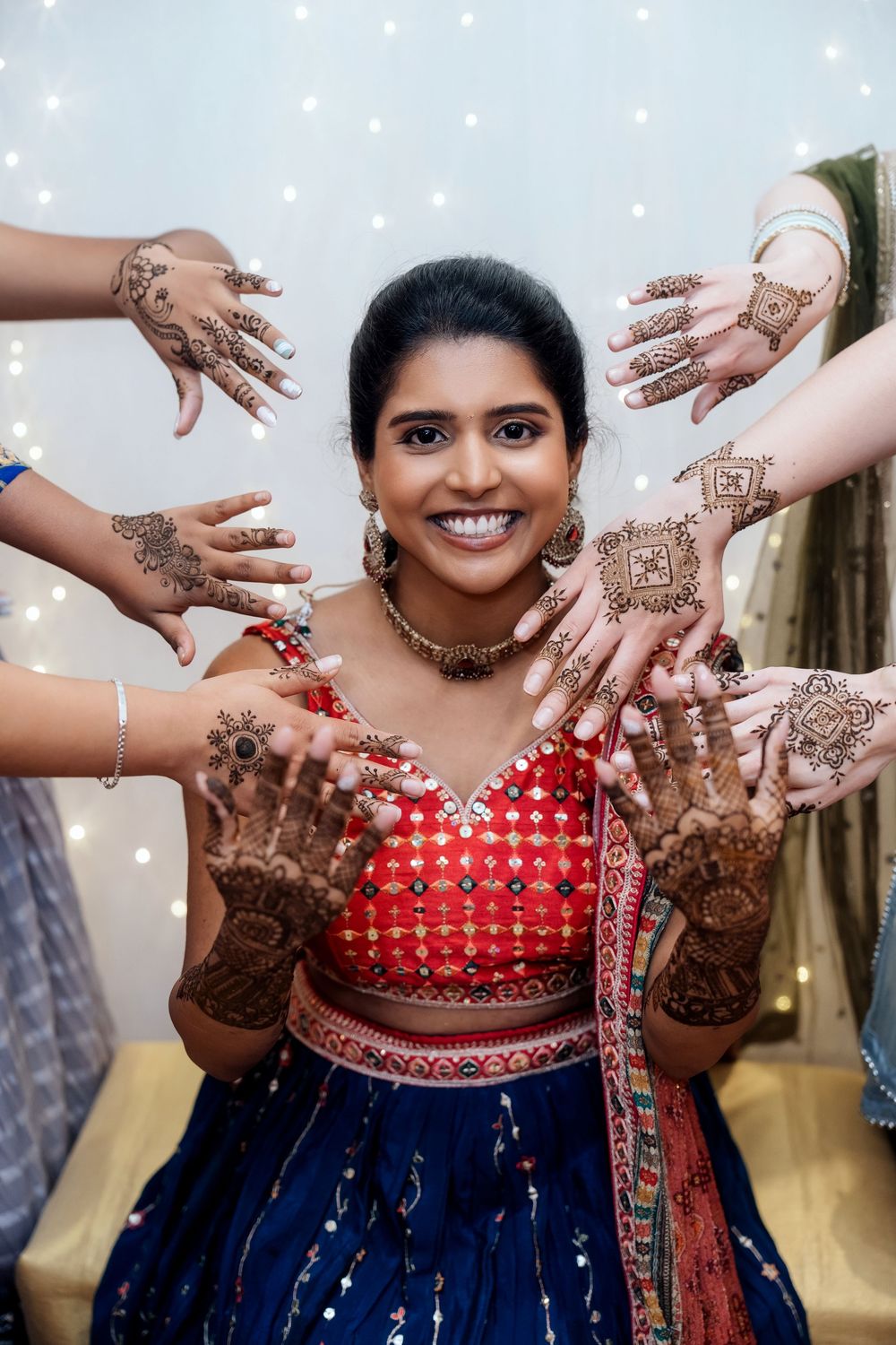 Multiple hands with henna designs surround a person in red and blue Indian wedding attire with traditional jewelry.