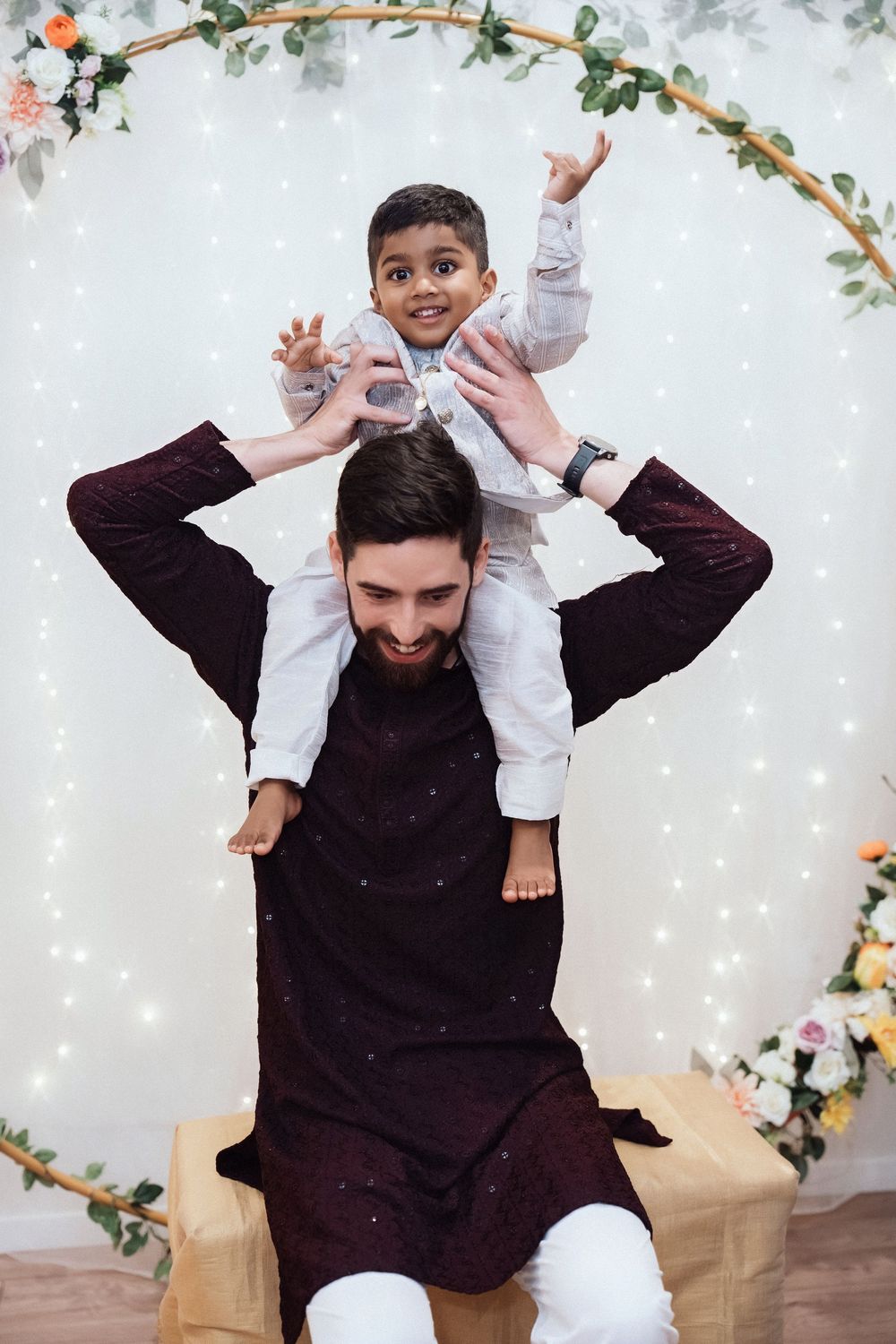 A person in a black outfit plays and interacts with a small child against a floral backdrop with twinkle lights.