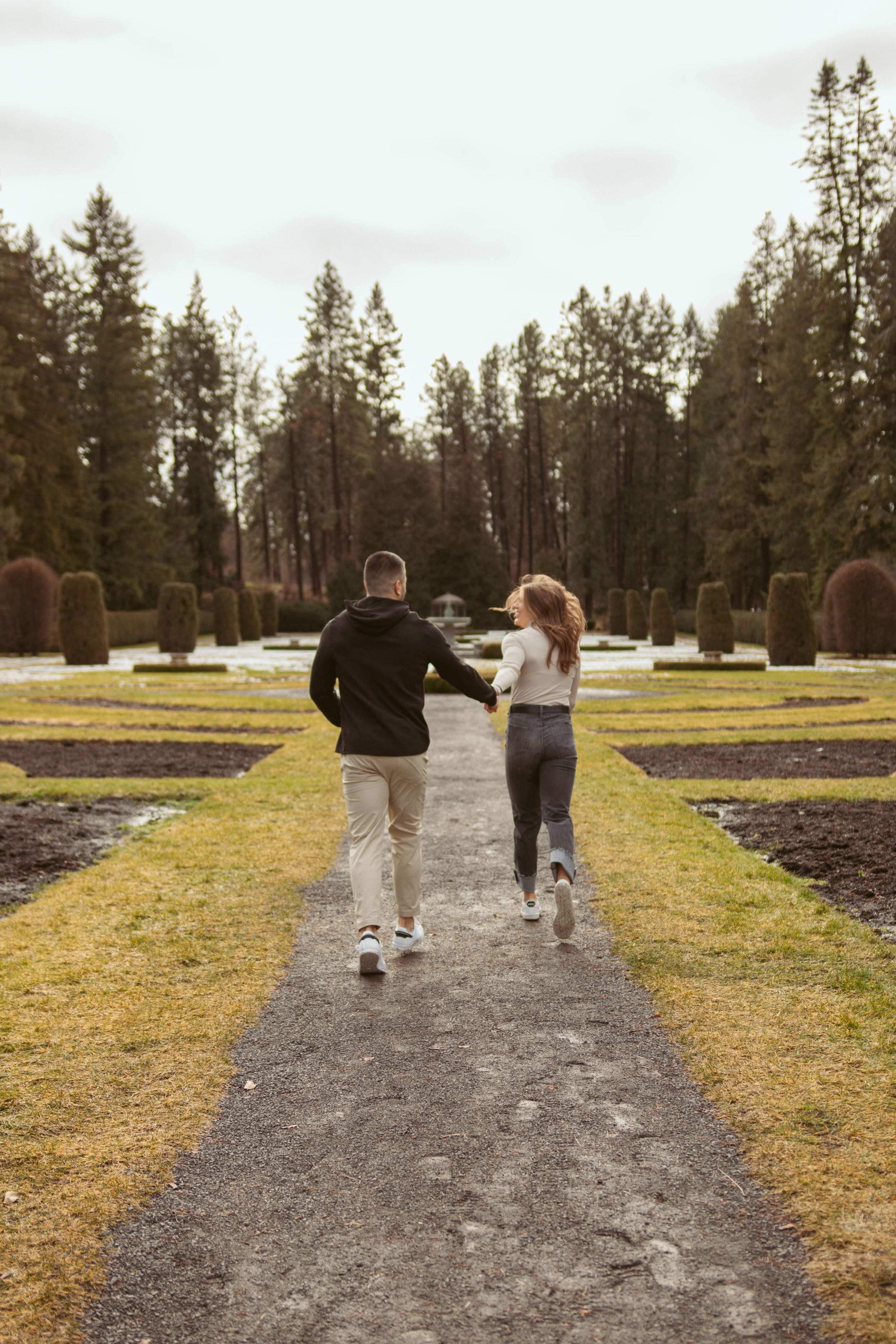 Two people running together on a path lined with evergreen trees and manicured gardens.