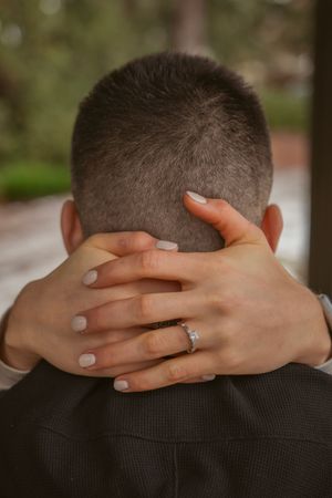 Two hands with light pink nails and an engagement ring clasp the back of a person wearing a dark suit.