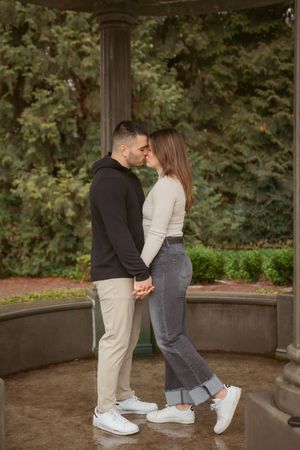 A couple in casual wear share an intimate moment while standing together in a gazebo surrounded by greenery.